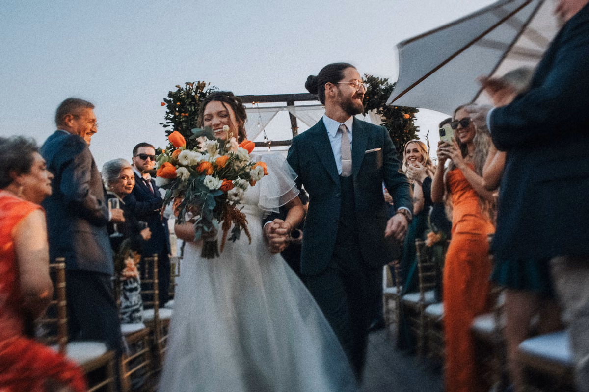 Novios salen sonrientes por el pasillo, ella con gran ramo blanco y naranja, invitados aplauden bajo una carpa.